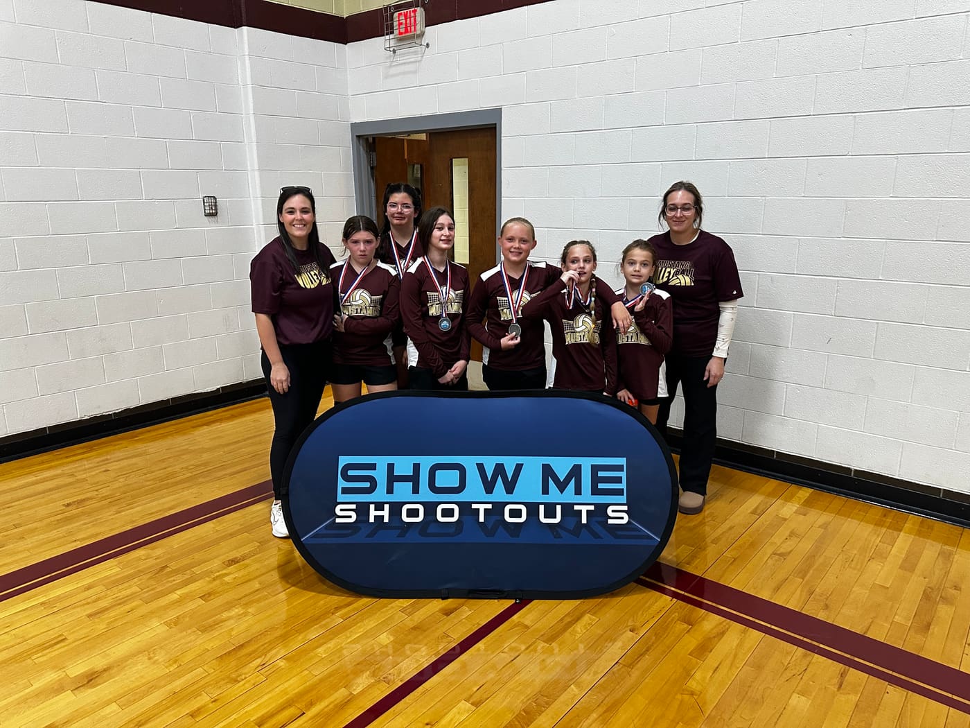 A Mustangs team in maroon jerseys shows off their medals alongside two coaches.