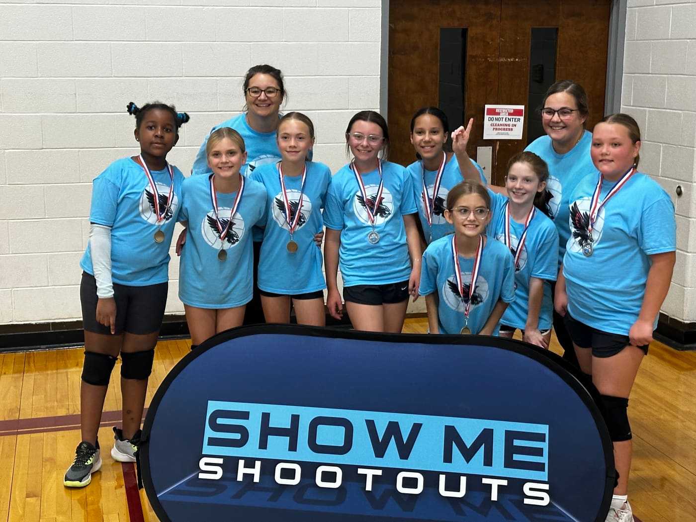 A team in light blue eagle shirts poses with medals and coaches, one player pointing a finger up for first place.