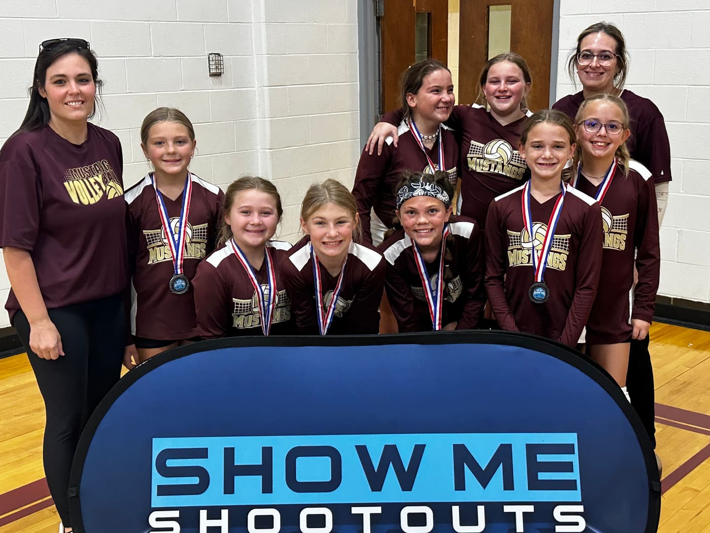 A Mustangs team in maroon jerseys smiles with their medals and two coaches behind the Show Me Shootouts banner.