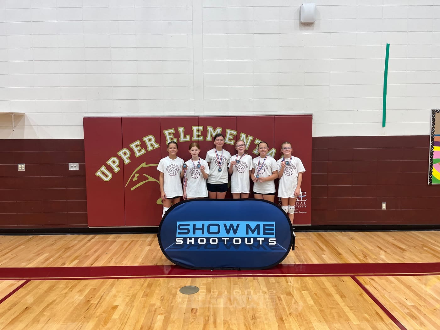 Six players in white Mustangs shirts hold their medals in front of the Upper Elementary wall pads.