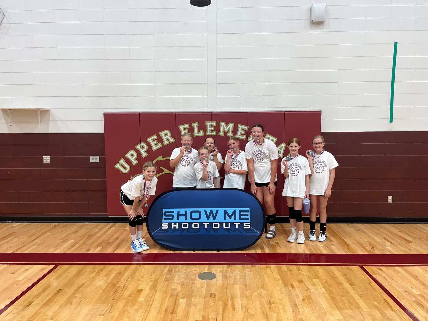Players in white Mustangs shirts bite their medals in front of the Upper Elementary wall pads.