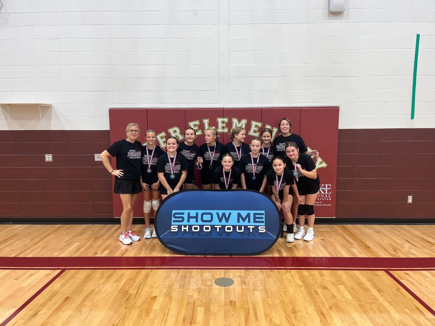 An Osage Indians team in black shirts poses with two coaches in front of the Upper Elementary gym wall.